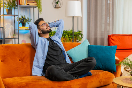 Happy relaxed young Indian man resting on couch with wireless earphones listening to ambient calming...