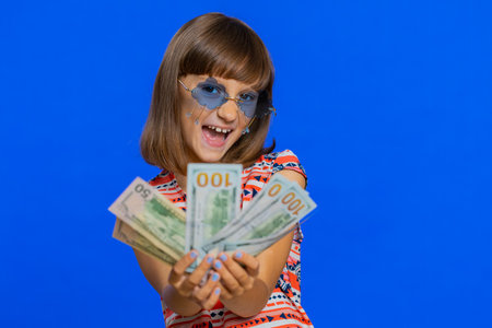 Rich pleased young cute school girl waving money dollar cash banknotes bills like a fan, success, lottery winner, big income, wealth. Teen brunette female child kid on studio blue backgroundの写真素材
