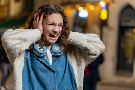Displeased angry young woman frowns face feels disgust suffering from loud sound covers ears with...