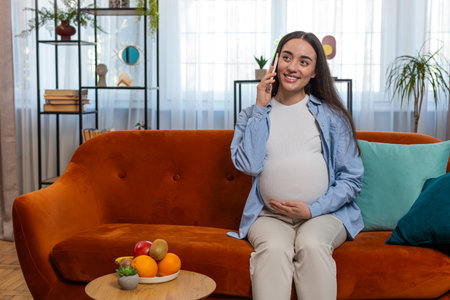Smiling pregnant woman talking on smartphone sitting on couch in living room at home. Happy Caucasian lady stroking belly holding cellphone in apartment. Maternity prenatal care and pregnancy concept.の写真素材
