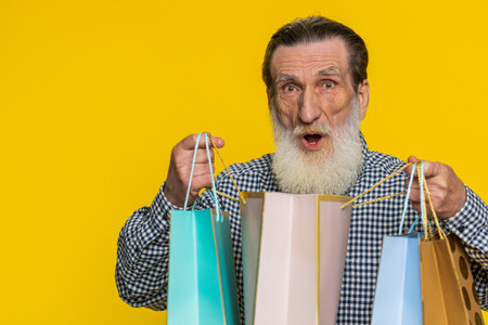 Happy amazed senior old man showing shopping bags advertising discounts smiling looking amazed with low prices shopping on Black Friday holiday sale. Elderly grandfather isolated on yellow backgroundの写真素材
