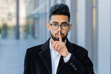 Shh be quiet please. Indian businessman presses index finger to lips makes silence hush gesture sign...