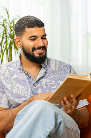 Indian man reading interesting book, turning pages smiling enjoying literature, taking a rest on comfortable couch. Peaceful cheerful guy relaxing at home apartment living room on sofa. Verticalの写真素材