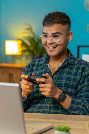 Excited Indian Arabian man playing video game on laptop spending leisure time at home office desk. Happy Hispanic freelancer guy playing video games with joystick controller on computer. Verticalの写真素材