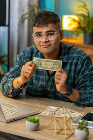 Indian man freelancer counting dollar cash insufficient amount of money. Financial crisis. Bankruptcy. Poverty and destitution. Hispanic male guy businessman with laptop sitting at home office tableの写真素材