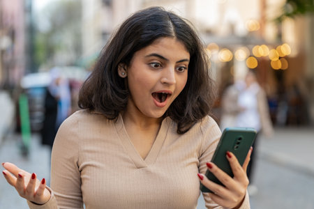 Oh my God Wow. Excited happy joyful winner Indian young woman use smartphone typing browsing celebrating win good message news outdoors. Hispanic girl walking on urban city street. Town lifestylesの写真素材