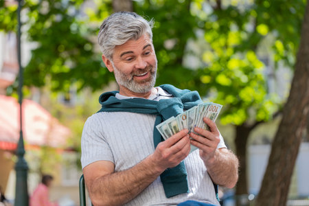 Rich happy mature man counting money cash, calculate domestic bills salary sitting on chair. Bearded guy satisfied of income earnings saves money for planned gifts. Bearded guy tourist on city streetの写真素材