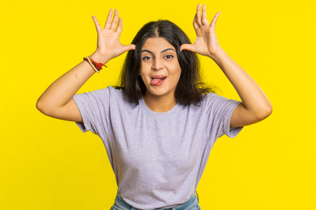Playful Indian young woman showing tongue making faces at camera, fooling around, joking, aping with silly face, teasing. Cheerful funny girl isolated on yellow backgroundの写真素材