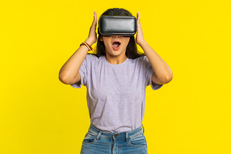 Excited happy young woman using headset helmet app to play simulation game. Watching virtual reality 3D 360 video. Girl in VR goggles isolated on yellow background. Future technologyの写真素材