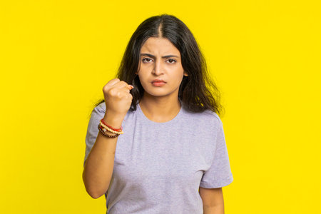 Angry Indian young woman trying to fight at camera, shaking fist, boxing with expression, mad fury. Girl isolated on yellow studio backgroundの写真素材