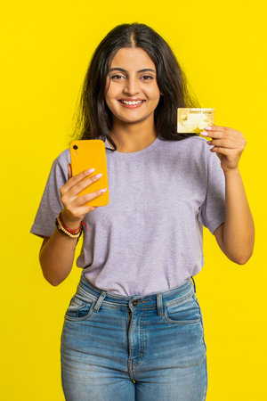 Happy Indian young woman using credit bank card and smartphone while transferring spending money,...