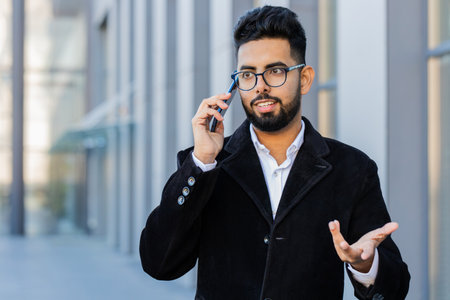 Stylish Indian businessman having remote conversation communicate speaking by smartphone outdoor. Arabian Hindu freelancer guy talking on phone standing downtown city street. Business people lifestyleの写真素材