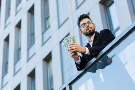 Happy rich Indian businessman winner counting money cash dollars banknotes salary celebrate victory, profit finance income reward outdoors. Arabian Hindu freelancer in downtown city. Business peopleの写真素材