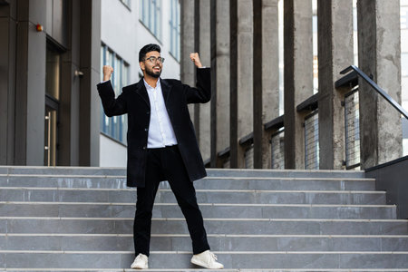 Happy Indian businessman runs down the stairs celebrating success, winning, career achievement, good news, luck, victory, successful signing of contract. Arabian Hindu freelancer guy in downtown cityの写真素材