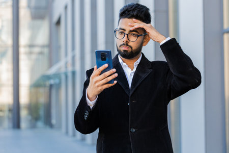 Indian businessman use smartphone typing browsing, loses surprised by bad news, fortune loss, fail bankruptcy outdoors. Arabian Hindu freelancer guy in downtown city street. Business people lifestyleの写真素材
