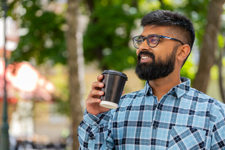 Happy Indian bearded man enjoying morning coffee hot drink and smiling outdoors. Relaxing, taking a break. Hispanic guy standing on urban city street drinking coffee to go. Town lifestyles. Horizontalの写真素材