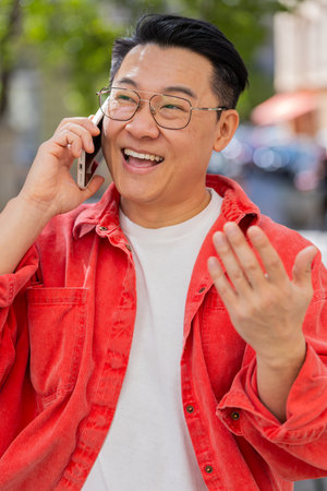 Happy Asian middle-aged man having remote conversation communicate speaking by smartphone outdoors....