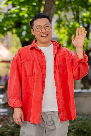 Hello. Asian middle-aged man smiling friendly at camera, waving hands gesturing hi, greeting or...
