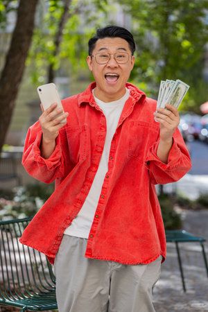 Happy rich Asian middle-aged man counting money dollar cash use smartphone calculator app plans to...