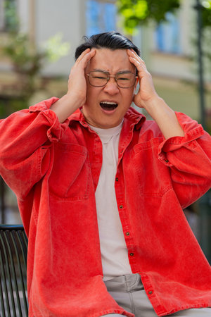 Sad unhappy Asian middle-aged man suffers from unfair situation outdoors. Problem, feeling bad, annoyed, burnout, grief. Frustrated depressed lonely Chinese guy sitting on urban city street. Verticalの写真素材