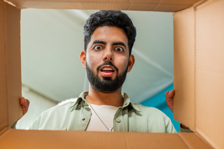 View from inside the box. Happy Indian man unpacking delivery parcel at home. Smiling satisfied guy shopper, online shop customer opening cardboard box receiving purchase gift by fast postal shippingの写真素材