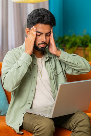 Tired Indian man freelancer use laptop, suffering from headache problem tension, migraine, stress at...