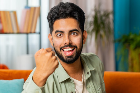 Happy Indian man shouting, celebrating success, winning, goal achievement good news, lottery luck, victory, watching TV championship at modern home apartment. Arabian guy in living room sits on couchの写真素材