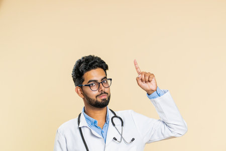Indian young doctor cardiologist man show thumbs up and pointing overhead empty place advertising area for commercial discount, copy space. Arabian apothecary pharmacy guy isolated on beige backgroundの写真素材