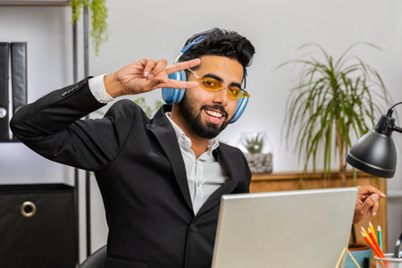 Happy relaxed overjoyed Indian businessman working on laptop computer at home office wearing headphones listening favorite energetic disco music and dancing. Freelancer man relaxing, taking a breakの写真素材