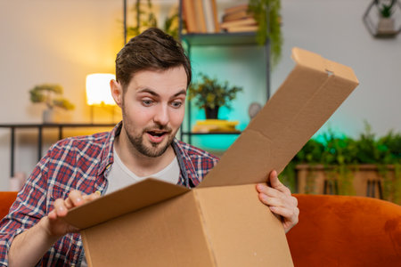 Happy excited man unpacking delivery parcel sitting on sofa in living room. Smiling Caucasian guy shopper online shop customer showing thumbs up receiving purchase by fast postal shipping courier.の写真素材