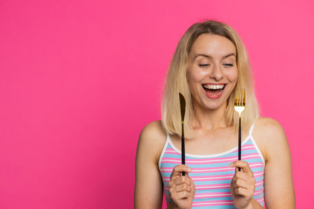 Ready to eat. Hungry Caucasian blonde woman in crop top waiting for serving dinner dishes with with restlessness holding cutlery fork knife will appreciate delicious restaurant meal on pink backgroundの写真素材