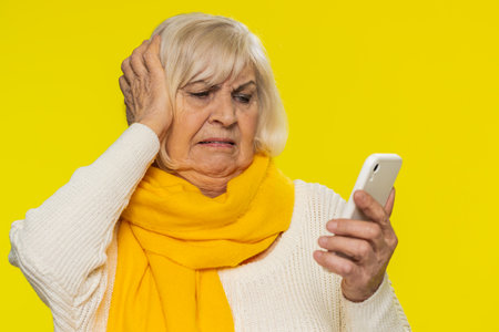 Upset sad senior woman use smartphone typing browsing, play game loses surprised by sudden lottery results bad fortune loss fail unlucky news. Elderly mature pensioner grandmother on yellow backgroundの写真素材