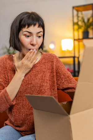 Happy excited young woman unpacking delivery parcel sitting at home. Smiling satisfied girl shopper...