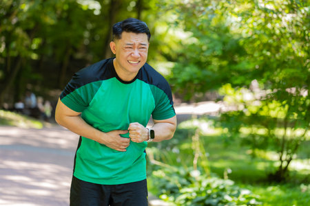 Middle-aged Asian runner feeling sudden strong abdominal stomach ache, gastritis problem outdoors in park. Man in sportswear having symptom poisoning diarrhea indigestion peptic ulcer pancreatitisの写真素材