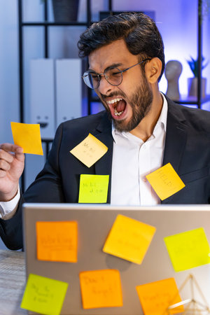 Tired exhausted Indian businessman working on laptop at office with many sticker task inscriptions....