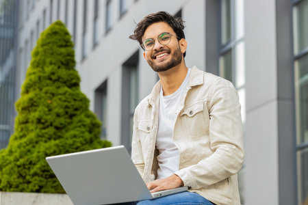 Happy young Indian man freelancer working on laptop computer sends messages, makes online purchases,...