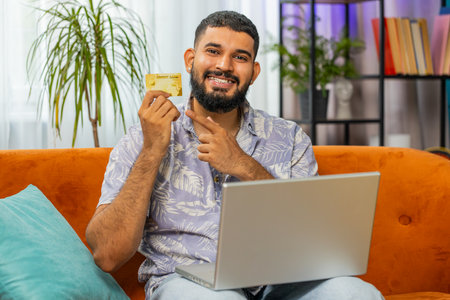 Indian man using credit bank plastic card and laptop computer while transferring money, purchases...