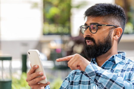 Indian bearded man using smartphone typing text messages in social media application online, surfing internet, relaxing, taking a break outdoors. Hispanic guy standing on urban city street. Horizontalの写真素材