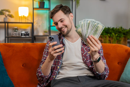 Successful rich man holding smartphone and waving dollar bill money fan sitting on sofa at home. Happy smiling Caucasian guy winning online casino lottery game planning vacation calculating budgetの写真素材