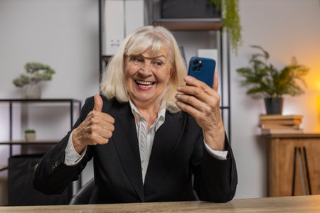 Happy mature woman using smartphone typing browsing yes celebrating success victory winning lottery jackpot goal achievement play game good positive news triumph. Elderly woman at home office table.の写真素材
