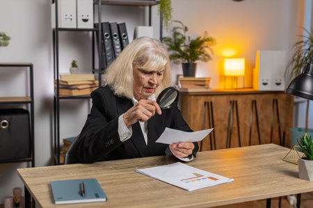 Accountant senior businesswoman looks through magnifying glass, investigate, examine analyzing reading documents, sits at workplace desk. Elderly grandmother freelancer working at office. Paperwork.の写真素材