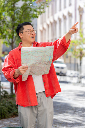 Middle-aged man tourist exploring city street, sightseeing holding paper map, checking direction searching way looking for the route outdoors. Travel concept. Town lifestyles. Verticalの写真素材