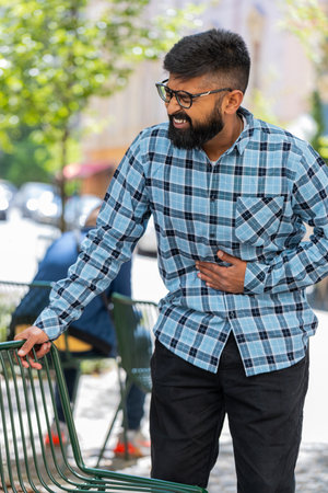 Young man feeling sudden strong abdominal stomach ache, gastritis problem outdoors. Guy having symptom poisoning diarrhea indigestion peptic ulcer pancreatitis on urban city streetの写真素材