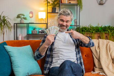 Amazed senior rich smiling man pensioner showing money dollar cash bills clenching at home room. Joyful happy mature middle-aged guy satisfied of income saves pointing money. Planning family budgetの写真素材