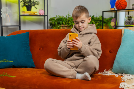 Young child boy texting share messages content on smartphone social media applications online watching relax movie. Male Caucasian teenager kid uses mobile phone at home in living room sits on sofaの写真素材