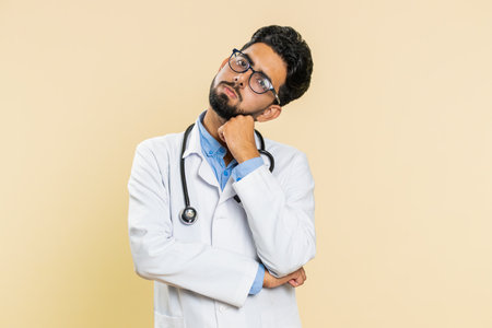 Sad Indian young doctor cardiologist man tired bored exhausted indifferent expression, not interested in communication talk, displeasure. Arabian apothecary pharmacy guy isolated on beige backgroundの写真素材