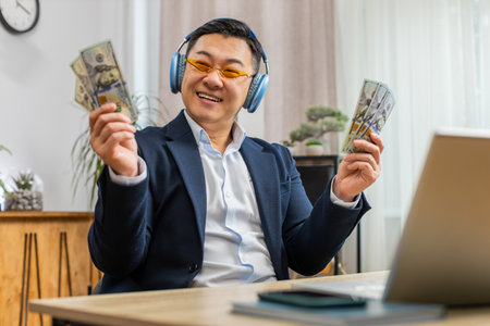 Asian business man holding fan of cash money dollar banknotes celebrate, dance, success career, lottery jackpot game winner, big income, wealth at home office workplace desk. Remote distant workingの写真素材