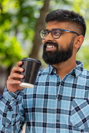 Happy Indian bearded man enjoying morning coffee hot drink and smiling outdoors. Relaxing, taking a break. Hispanic guy walking on urban city street drinking coffee to go. Town lifestyles. Verticalの写真素材