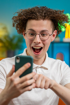 Surprised excited Caucasian young man winner holding smartphone reading good news amazed by online bet bid game win. Happy guy male on sofa looking at screen overjoyed by victory success in apartment.の写真素材