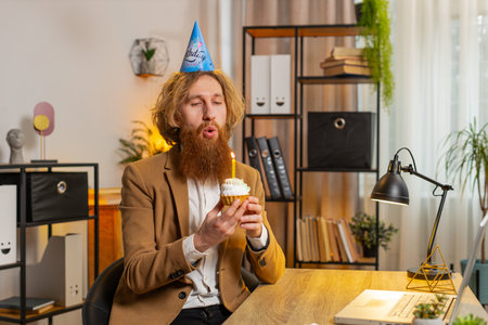 Happy Caucasian businessman wears festive birthday hat hold cupcake makes wish joyful congratulating blowing burning candle on cake. Redhead man freelancer celebrating birthday party at office aloneの写真素材
